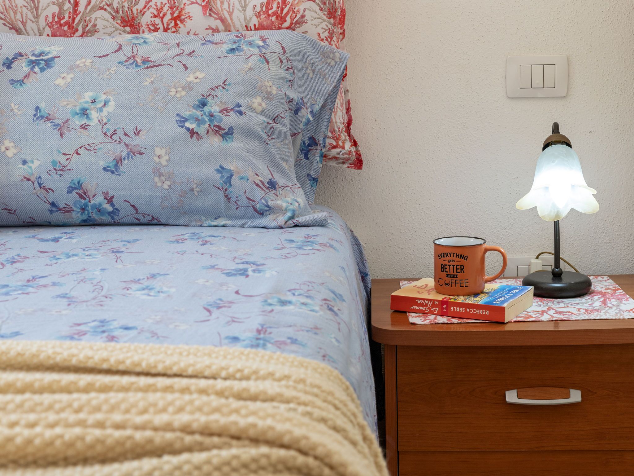 Un lit bien fait, avec une literie à motifs floraux, une table de chevet sur laquelle se trouvent une lampe, une tasse et des livres.