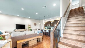 A modern living room with a wooden staircase, a flat-screen TV, and a kitchen area with bar stools. - 30A SANTA ROSA BEACH RETREAT EASY 5 MIN WALK - 4BR 5BA HOUSE W/POOL (Santa Rosa Beach)