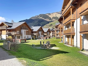 A row of wooden chalet-style buildings with balconies, a grassy courtyard, and a mountainous backdrop.