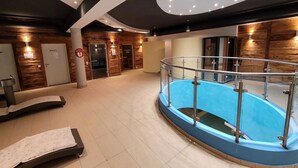 A modern indoor swimming pool with a glass railing, wooden walls, and a ceiling with recessed lighting.
