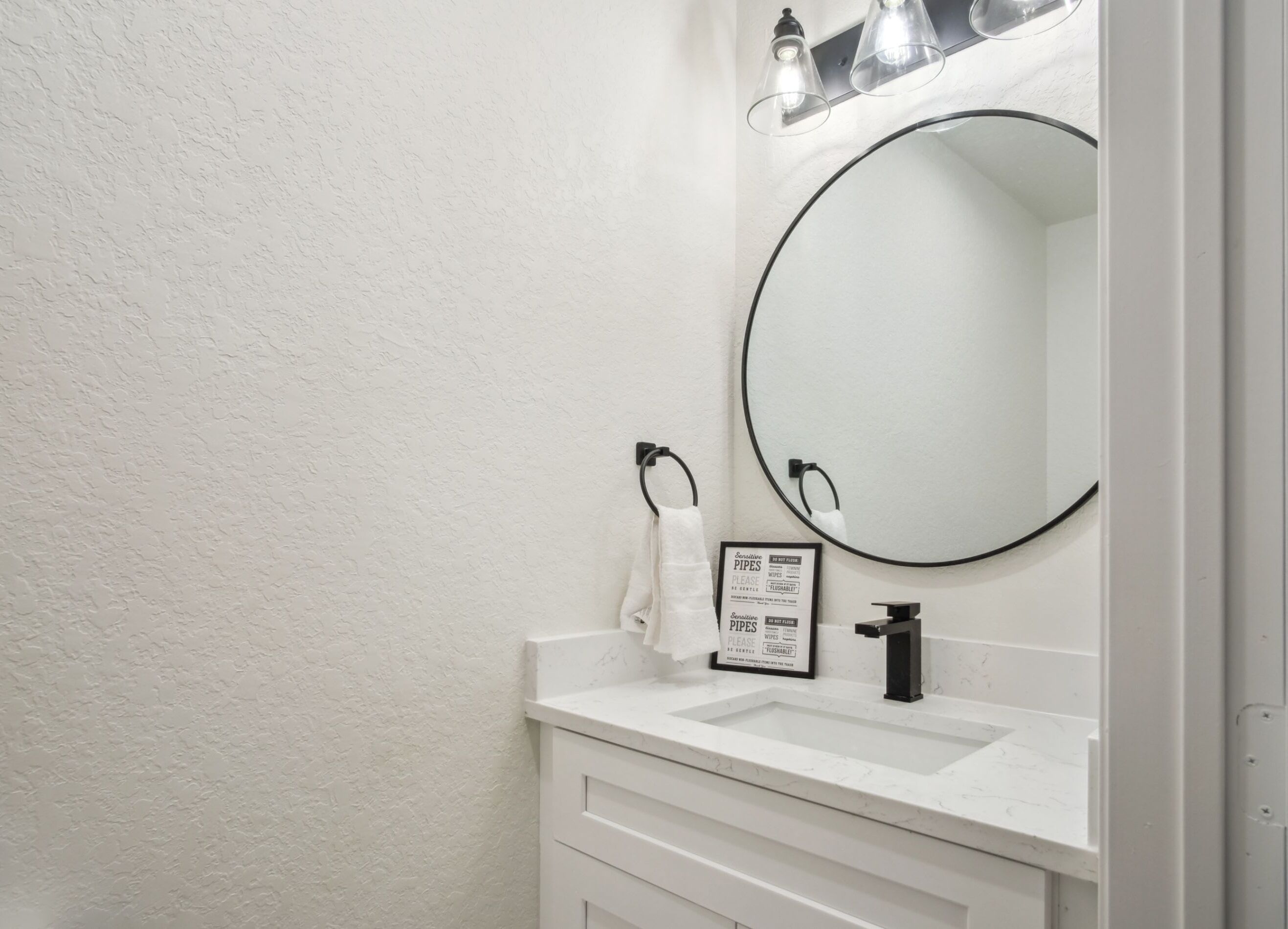 Un bagno con un lavandino bianco, uno specchio e un asciugamano appeso a un anello. Sullo specchio è presente un cartello e una lampada sopra di esso.