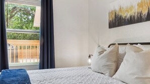 Une chambre d’hôtel avec un lit, du linge de lit blanc, un tableau au mur et une vue sur un balcon bordé d’arbres.