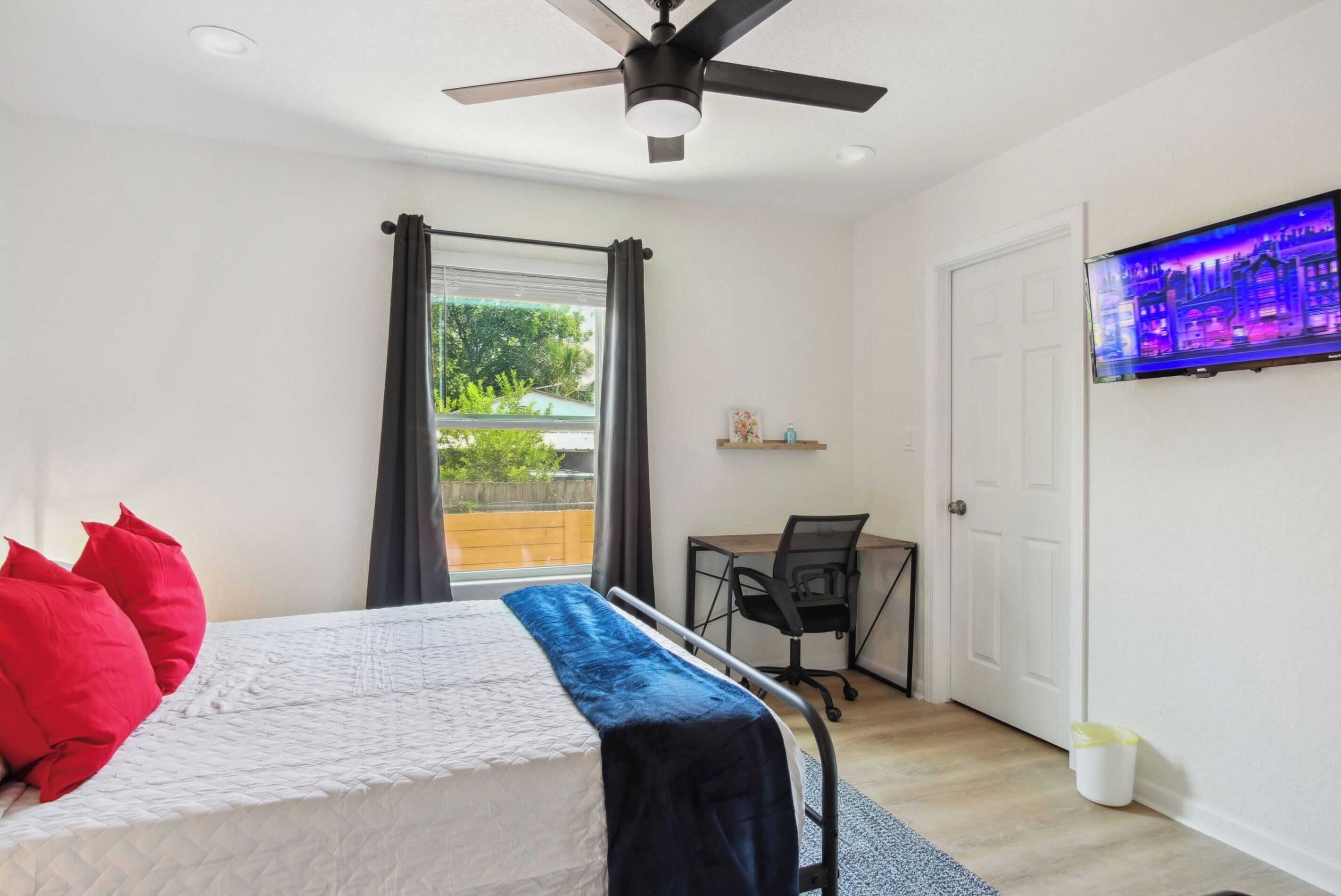 A bedroom with a bed, a desk, a chair, a ceiling fan, and a TV on the wall.