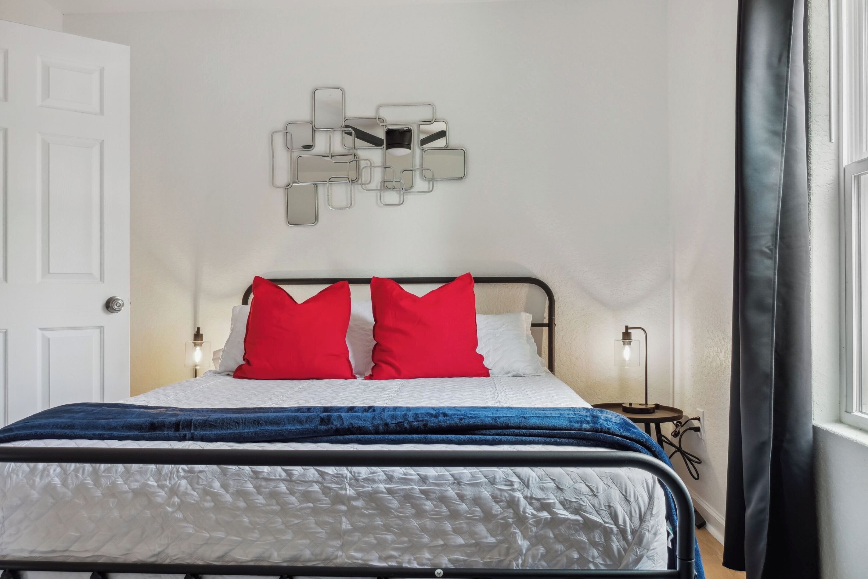 A neatly made bed with a white and blue quilt, two red pillows, and a wall-mounted abstract art piece.