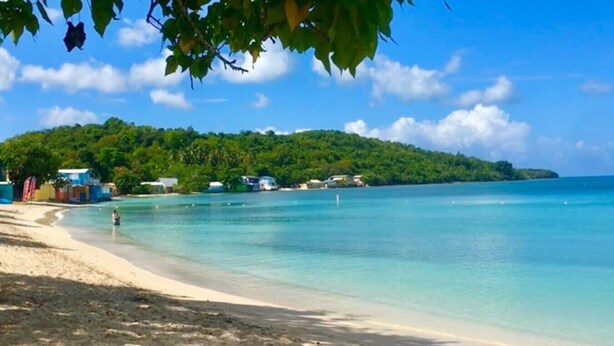 A beach with clear turquoise water, a sandy shore, and a small hill with buildings in the background. - Peaceful Hideaway in Cabo Rojo (Boquerón)