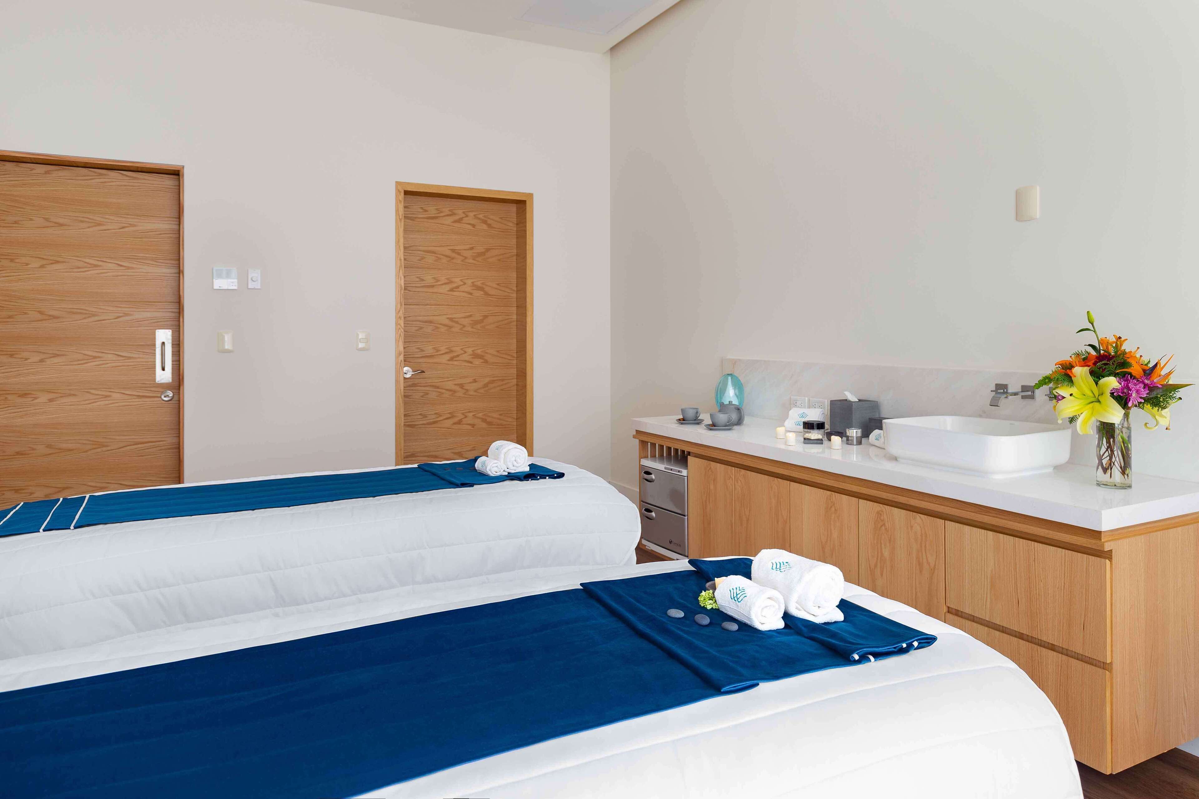Habitación de hotel con dos camas, una puerta de madera, un lavamanos blanco y un jarrón con flores.
