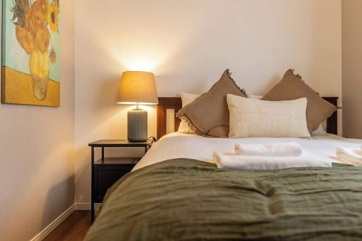 A neatly made bed with a green blanket, a bedside lamp, and two pillows on a nightstand.