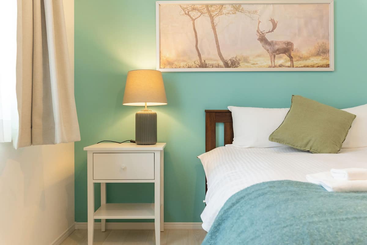 A bedroom with a bed, a nightstand with a lamp, a green wall, and a framed picture of a deer.