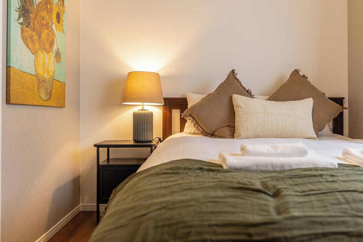 A hotel room with a bed, pillows, a lamp, and a painting on the wall.