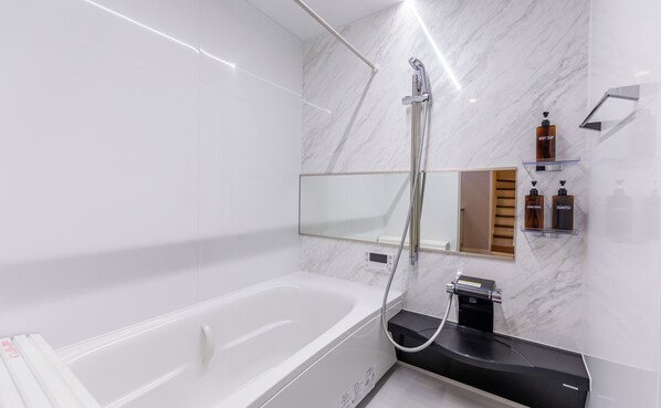 A modern bathroom with a white bathtub, a handheld showerhead, a built-in toilet, and a mirror above the sink. - es Maison Kasuga (Osaka)