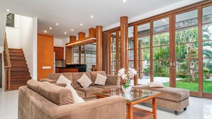 Amplia sala de estar con un ventanal grande, una escalera de madera y una cocina moderna al fondo.