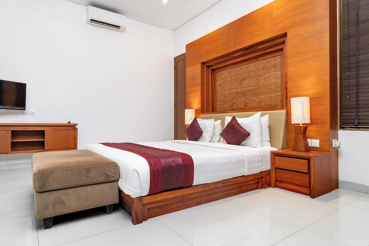 A modern bedroom with a wooden headboard, a large bed with white and red bedding, a wooden nightstand with a lamp, and a TV on a wooden stand.