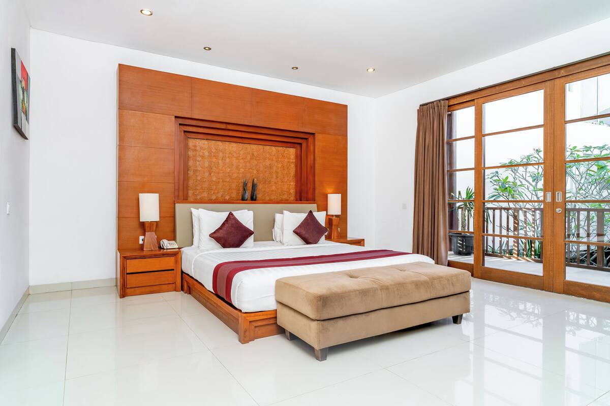A spacious bedroom with a large bed, a wooden headboard, a bedside table with a lamp, and a view of greenery through a sliding glass door.