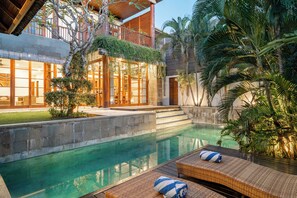 A poolside area with a lounge chair, a towel, and a view of a building with a balcony and a waterfall. - Bale Gede Luxury Villas by Moria (Seminyak)