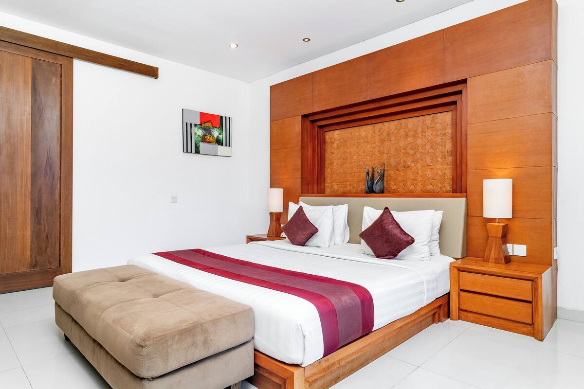 A modern bedroom with a wooden headboard, a large bed with a red and white bedspread, two bedside lamps, and a wooden nightstand.