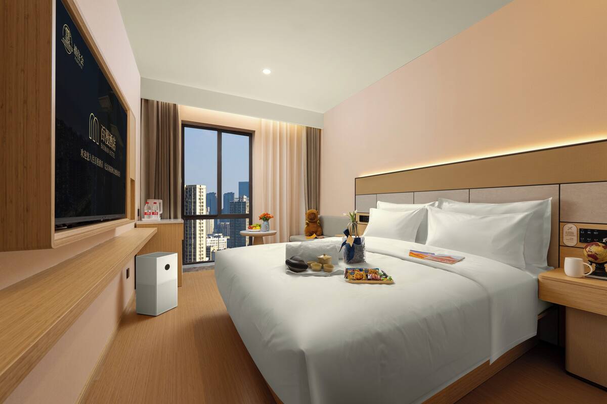 A hotel room with a large bed, a bedside table with a lamp, a window with a view of buildings, and a television on the wall.