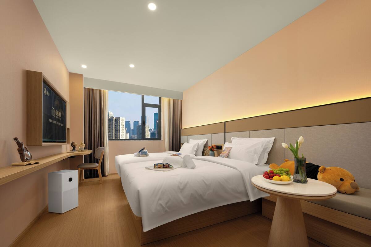 A modern hotel room with a large bed, a bedside table with a teddy bear and flowers, a desk with a computer, and a city view.