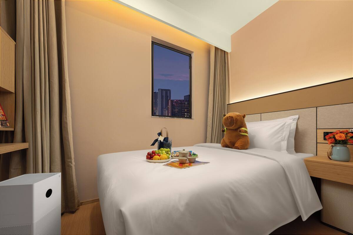A hotel room with a bed, a teddy bear, a window with a city view, and a bedside table with a vase of flowers.