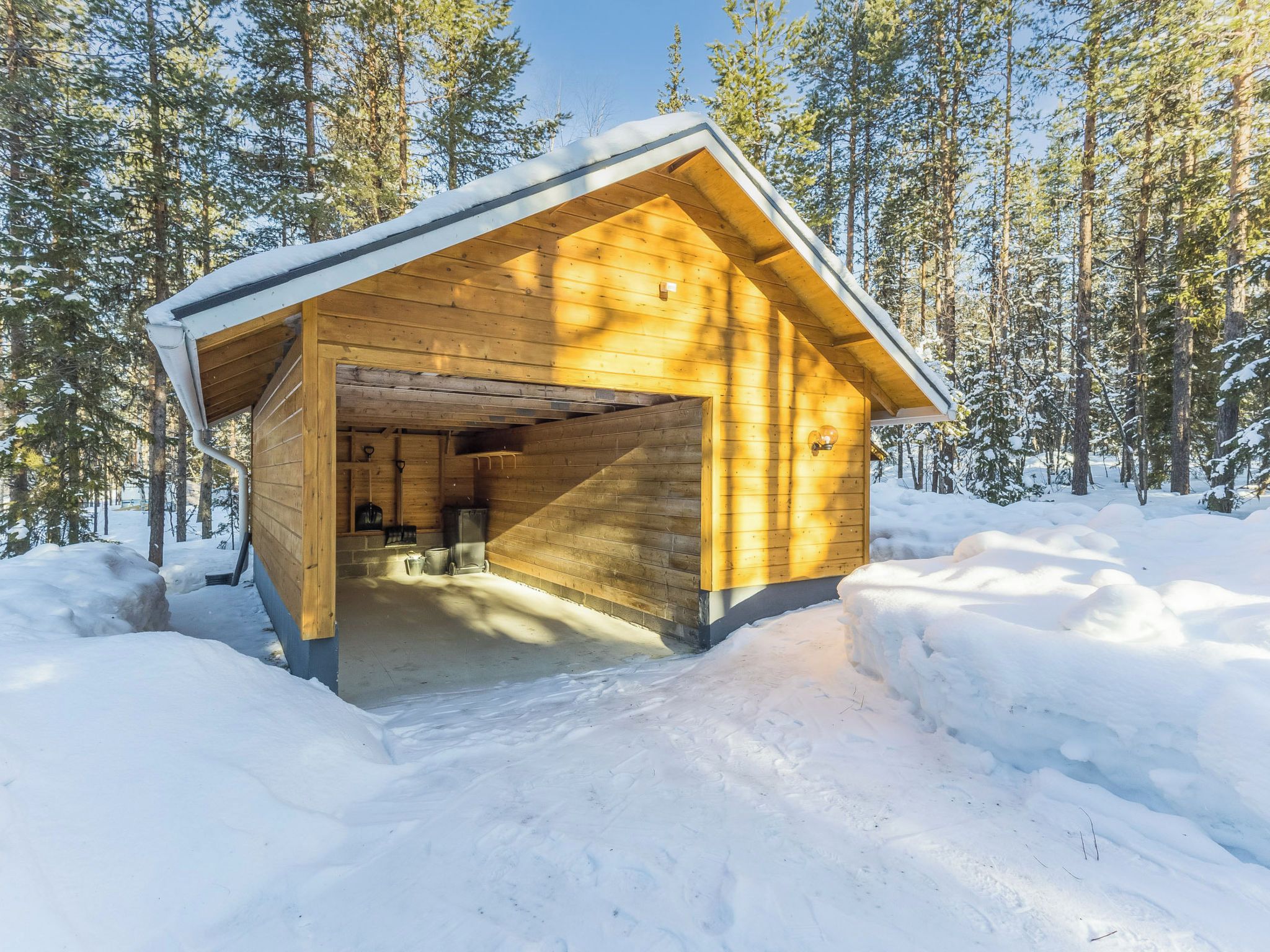 A wooden cabin with an open entrance, surrounded by snow-covered ground and trees.