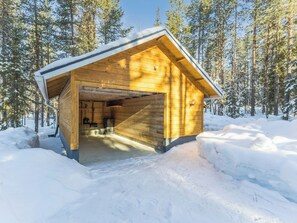 A wooden cabin with an open entrance, surrounded by snow-covered ground and trees.