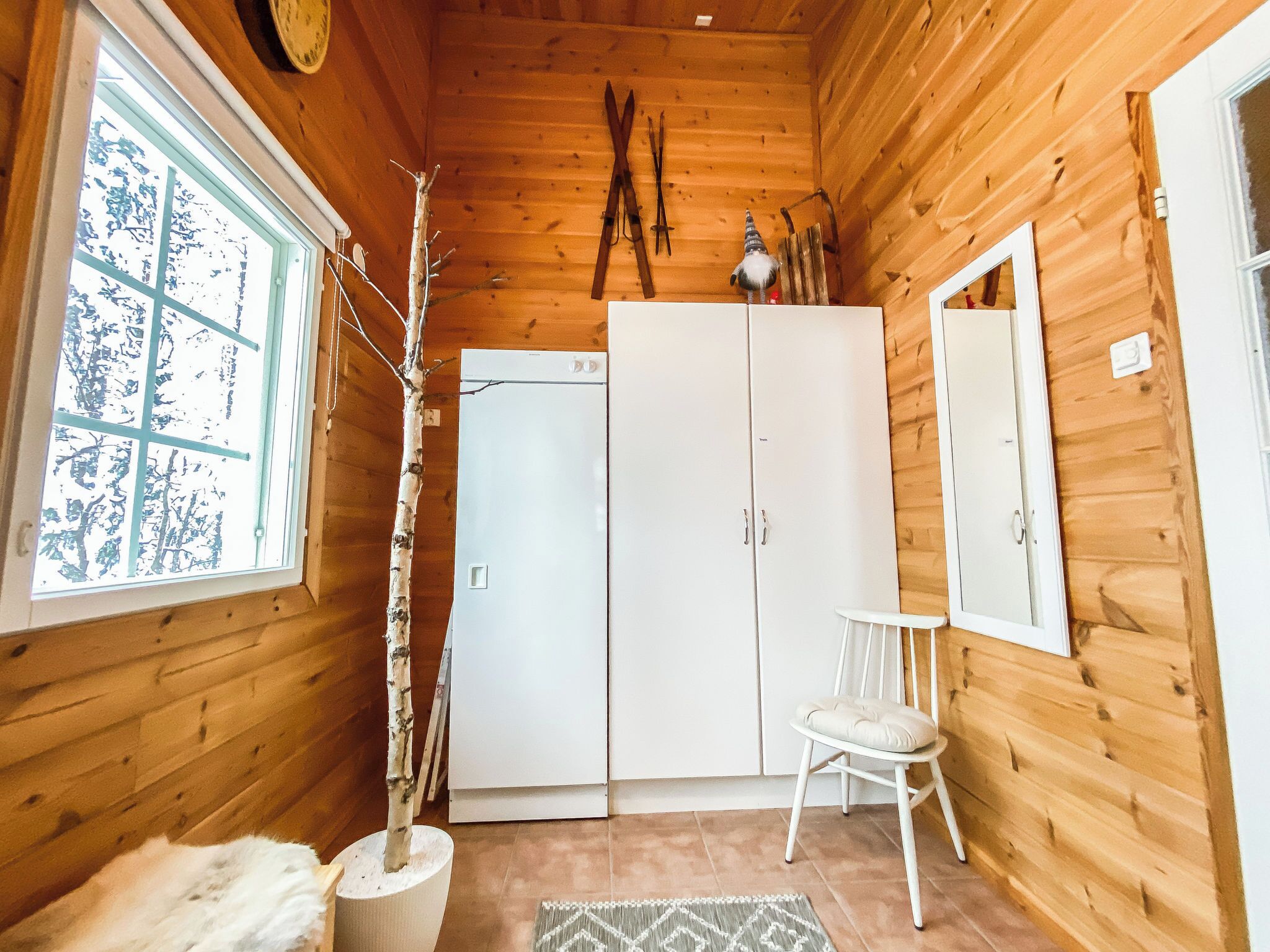 Interior de una cabaña de madera con un armario grande, una mesita, una silla y una ventana que da a vistas de árboles.