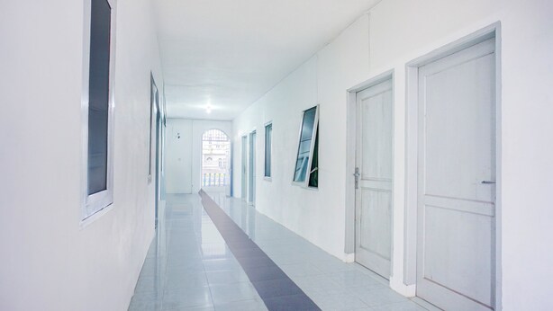 Un pasillo largo y estrecho con paredes y puertas blancas, que conduce a una habitación luminosa con un ventanal grande.