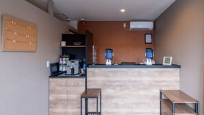 Un'area reception con un bancone in legno, una macchina da caffè e uno schermo digitale. Sono presenti due tavoli in legno e una bacheca a muro.
