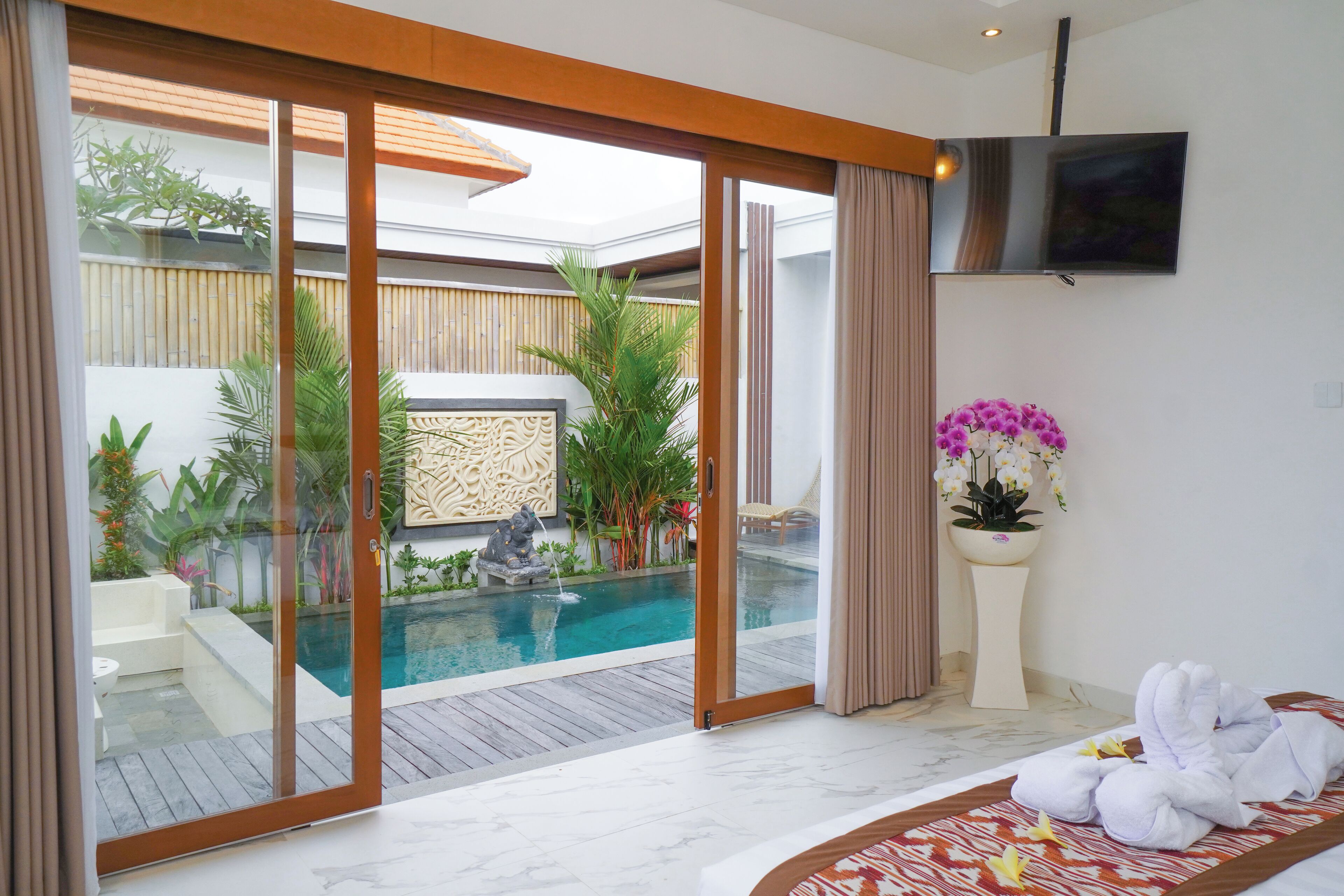 A modern hotel room with a large sliding glass door leading to a private pool area, a flat-screen TV mounted on the wall, and a potted orchid on a pedestal.
