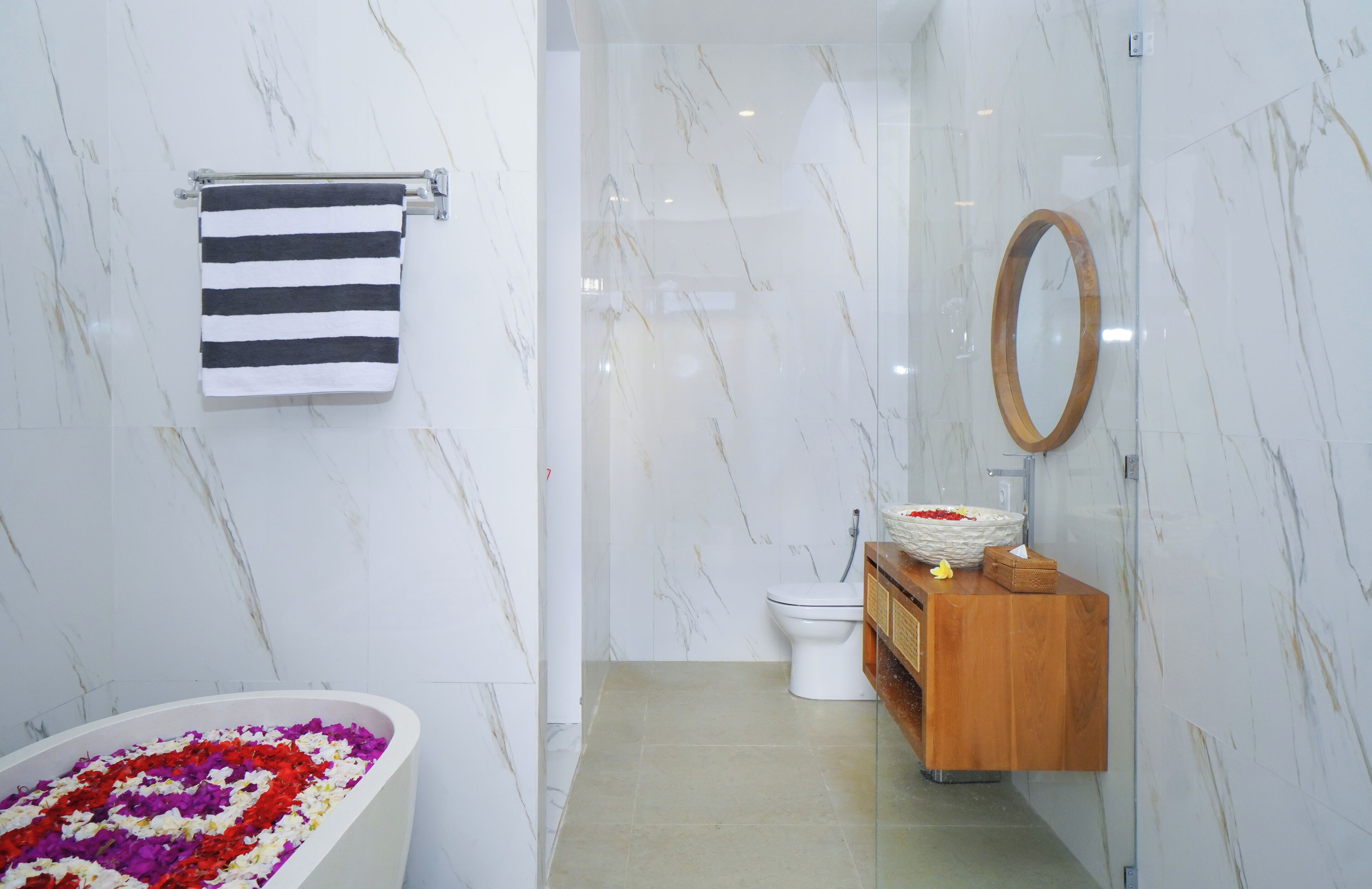 A modern bathroom with a marble wall, a striped towel, a wooden vanity, a toilet, and a bathtub with flower petals.