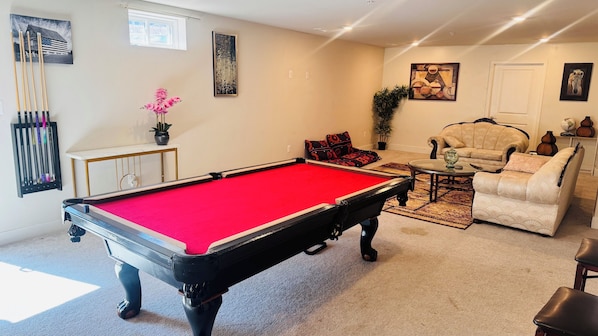 A pool table in a living room with a sofa and armchairs, a small table with a vase, and a potted plant.