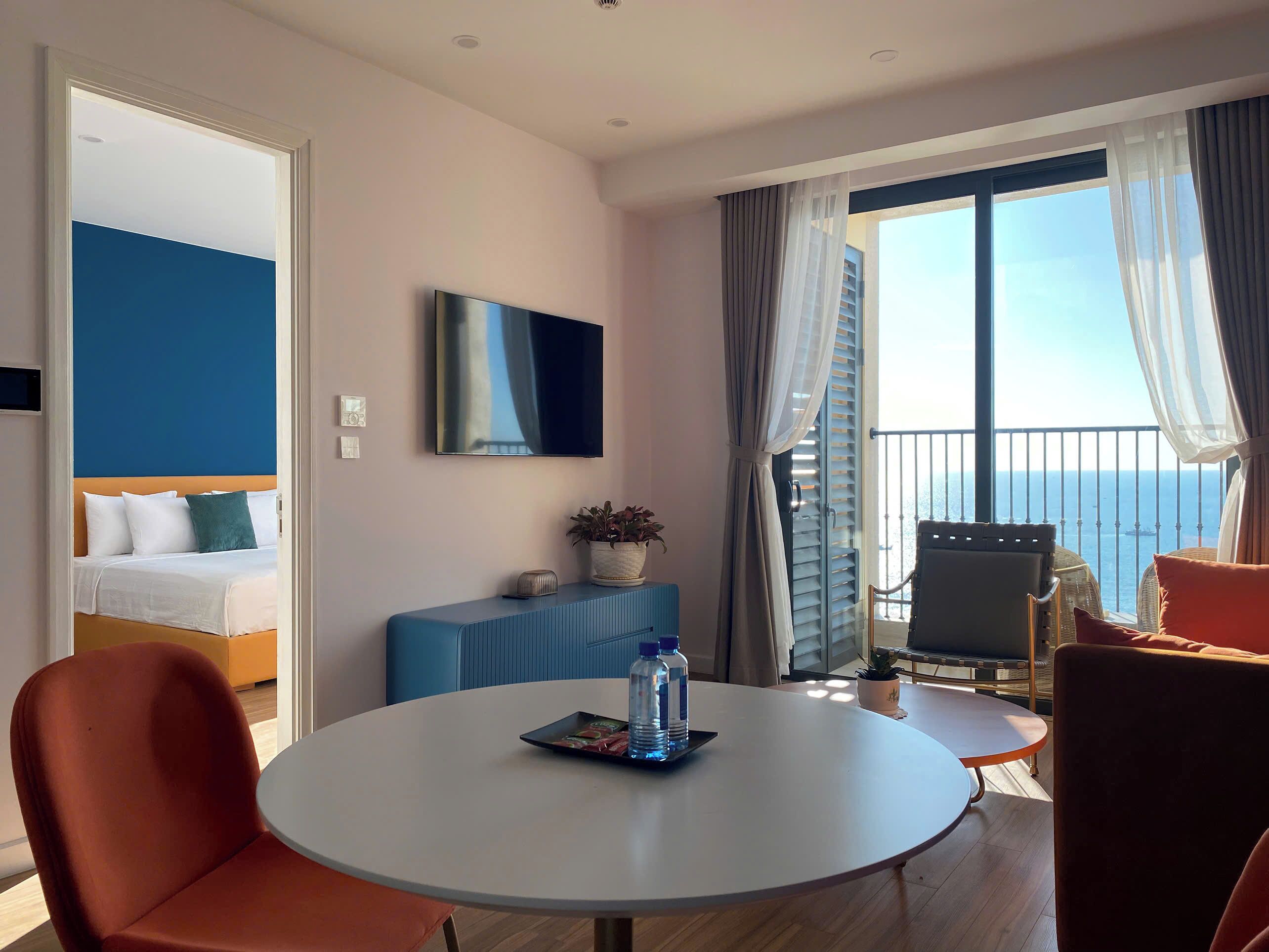 Una habitación de hotel moderna con una cama, una mesa redonda con sillas, un televisor y un balcón con vistas al mar.