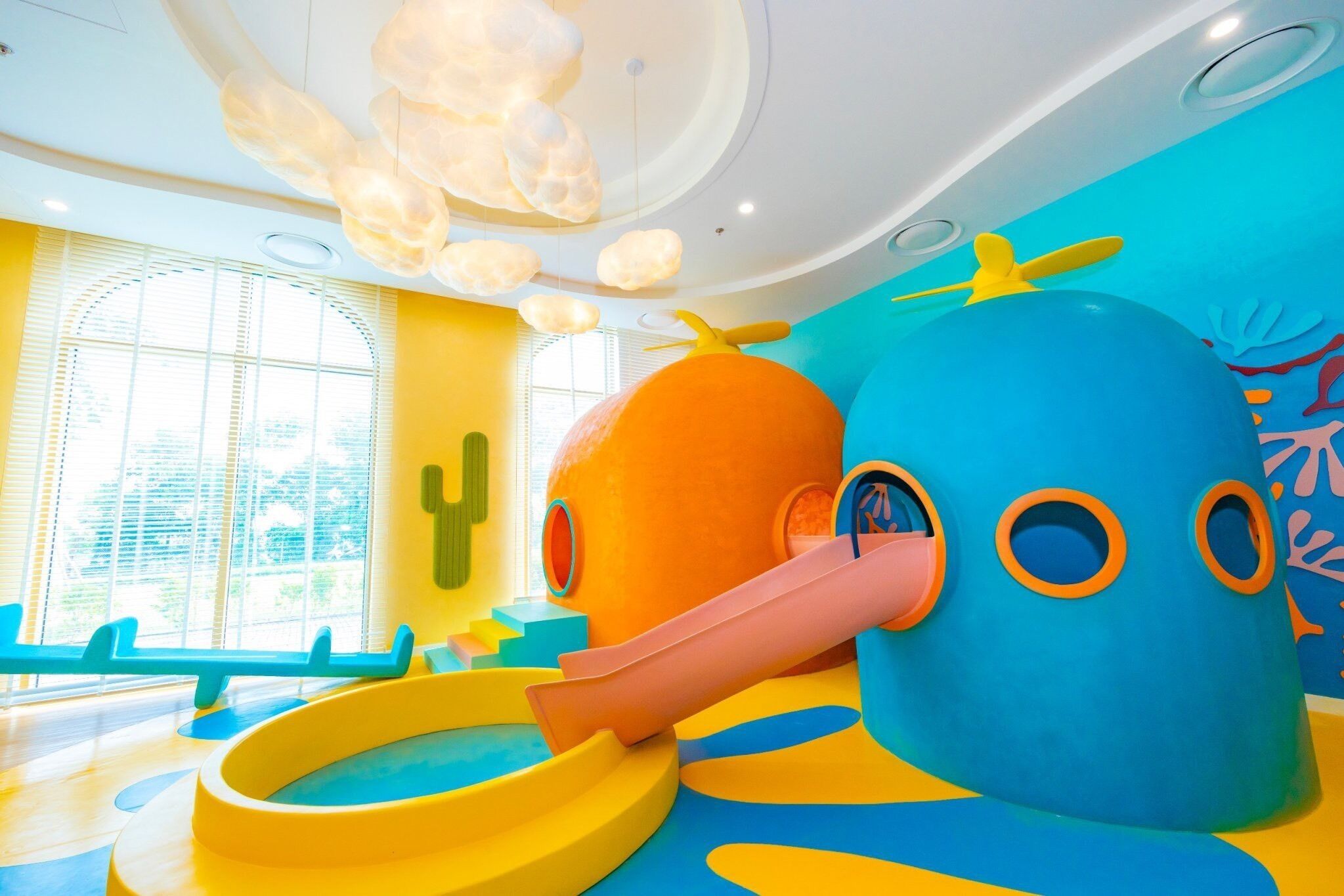 A colorful indoor play area with a slide, a large blue and orange structure, and a window with blinds.