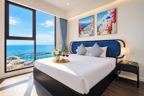 A hotel room with a large window, a bed with white linens, two pillows, a bedside table with a lamp, and two paintings on the wall. - Skyline Hillside Apartment Phu Quoc (Phu Quoc)