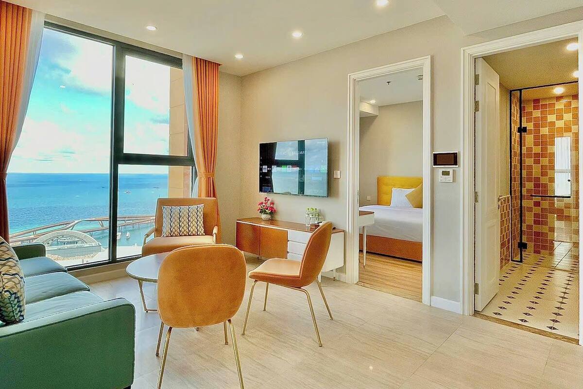 A modern hotel room with a large window offering a view of the ocean, a dining area with a table and chairs, a bed with a nightstand, and a bathroom with a tiled wall.
