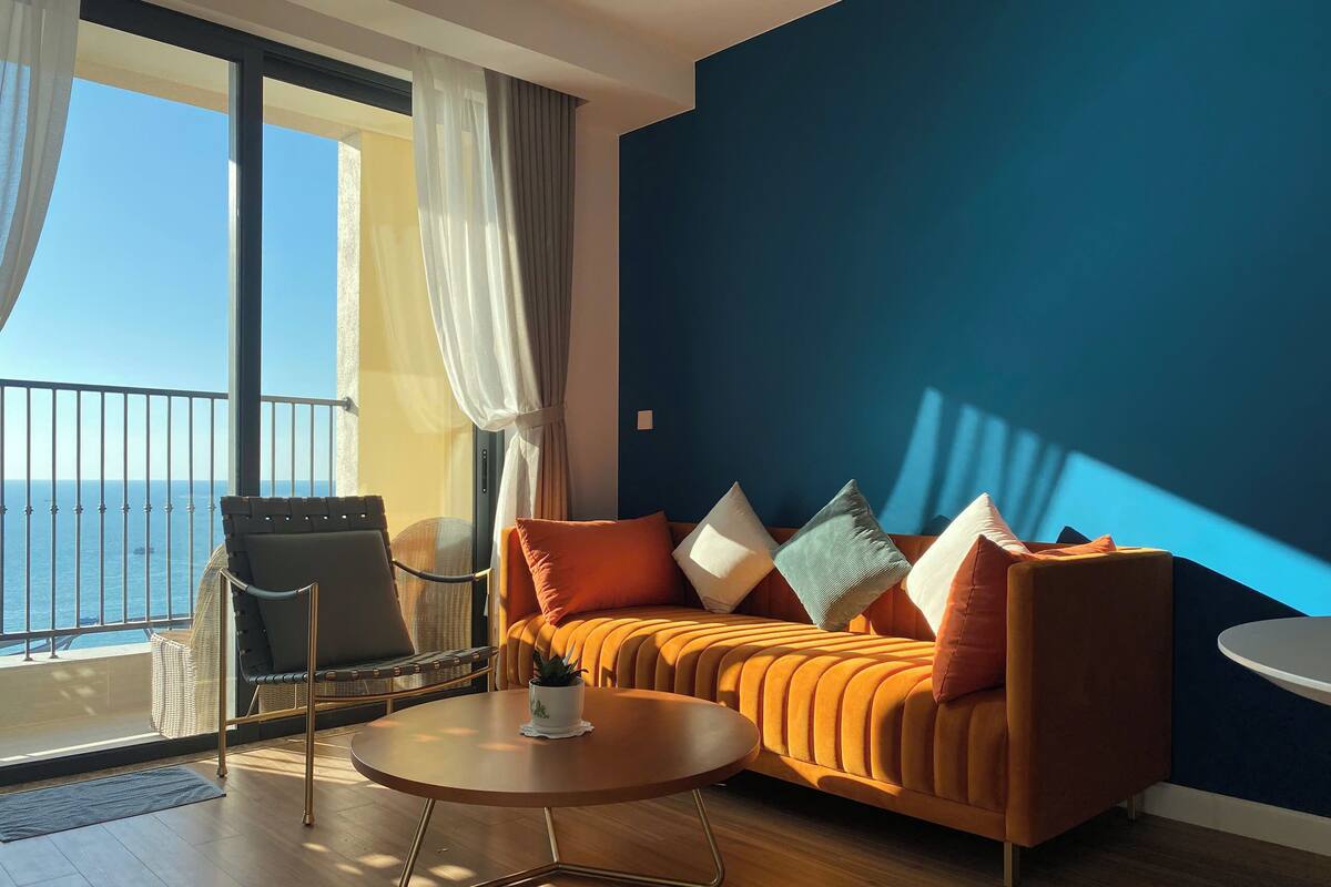 A modern living room with a sofa, a round coffee table, and a chair. There is a balcony with a view of the sea.