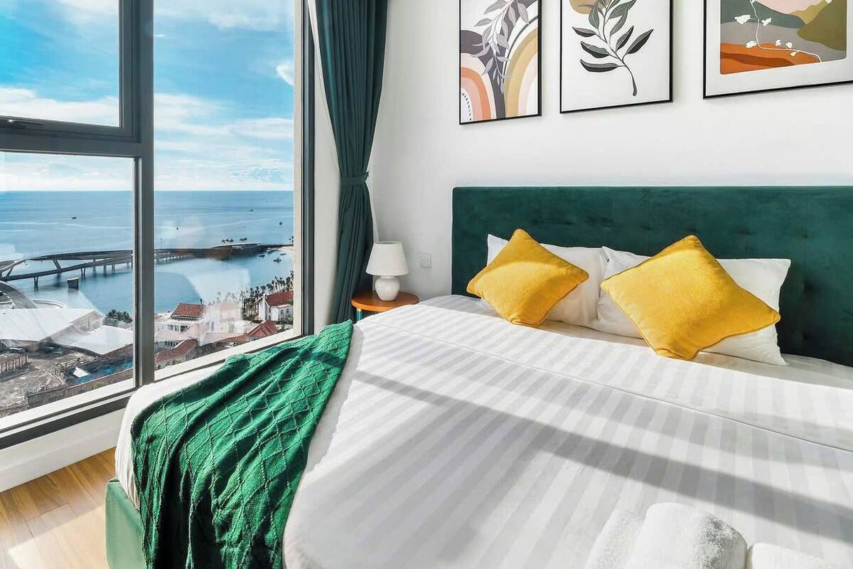A hotel room with a large window overlooking a coastal view, a bed with white bedding and two yellow pillows, and a green headboard.