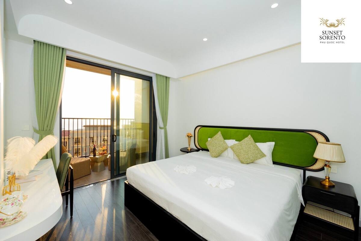 A hotel room with a large bed, a nightstand, a desk with a lamp, and a view of the city through a glass door.