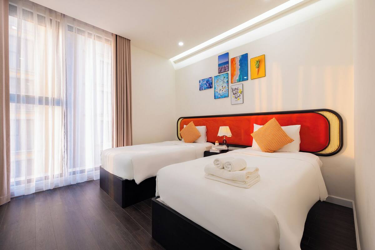 A hotel room with two beds, a large window with sheer curtains, a wall with framed artwork, and a modern headboard.