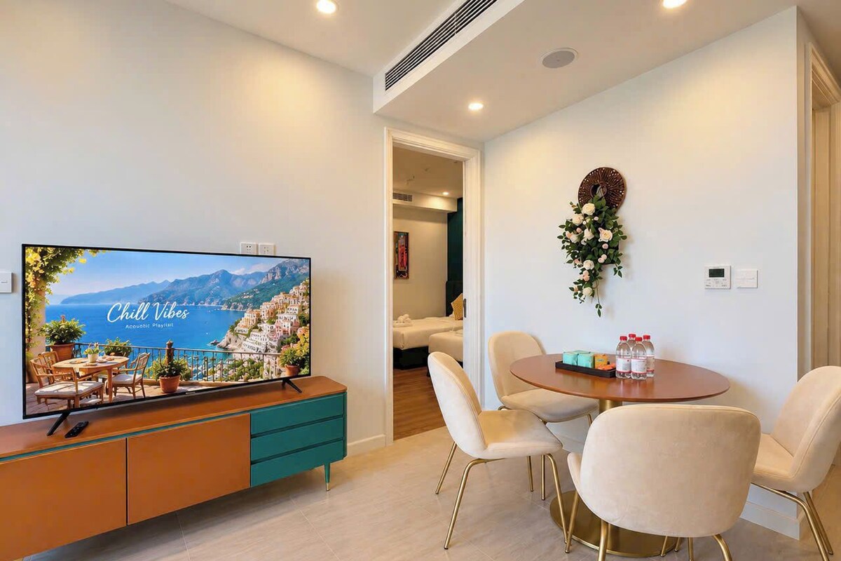 A modern living room with a flat-screen TV, a dining area with a round table and chairs, and a small balcony with a view.