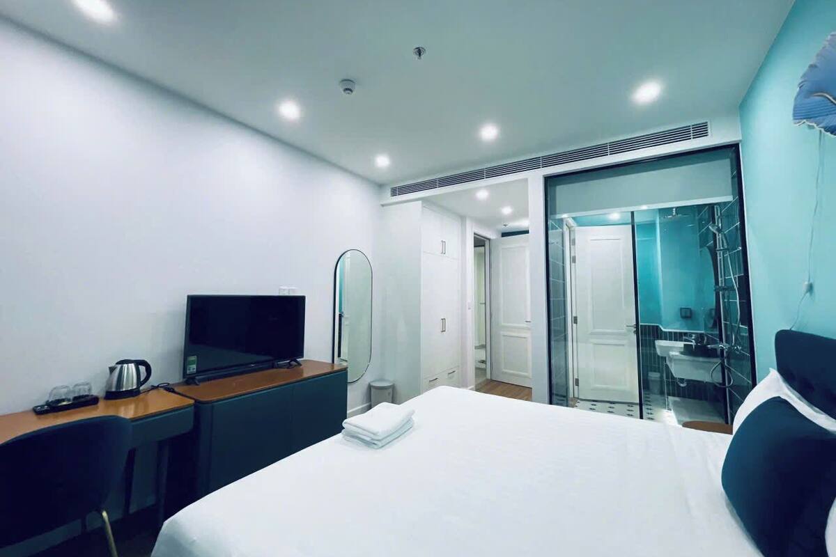 A modern hotel room with a bed, a desk with a television, a mirror, and a bathroom visible through a glass door.