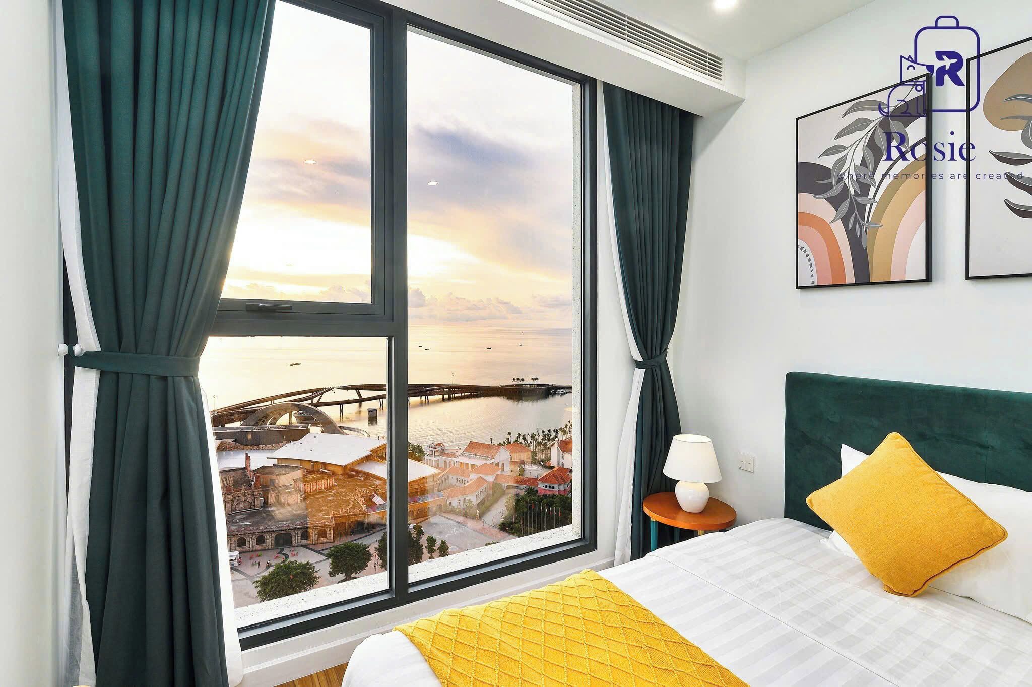 A hotel room with a large window, a bed with a yellow pillow, and a nightstand with a lamp.
