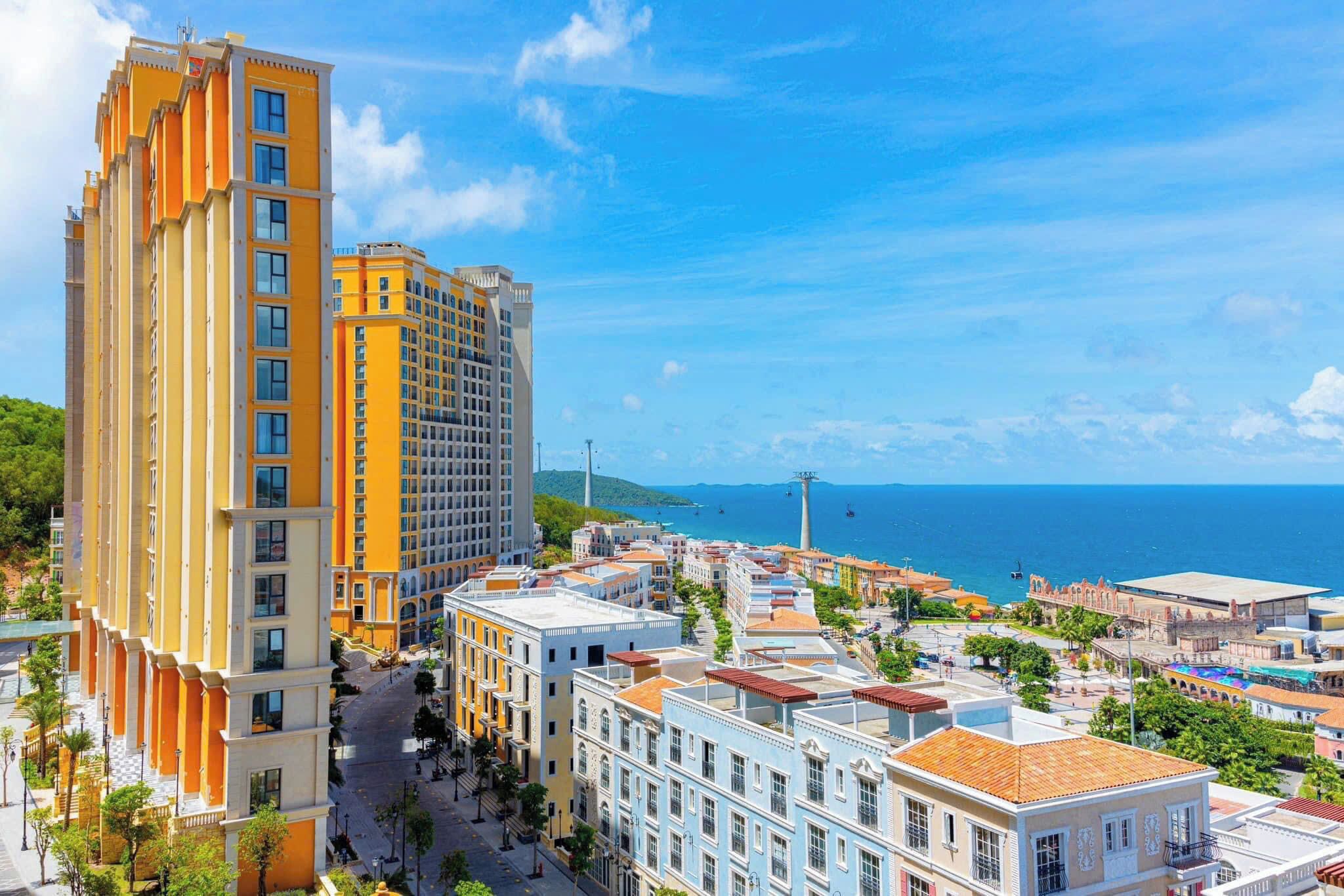Eine Küstenstadtansicht mit einem markanten gelben Hotel, verschiedenen farbenfrohen Gebäuden und freiem Blick auf das Meer.