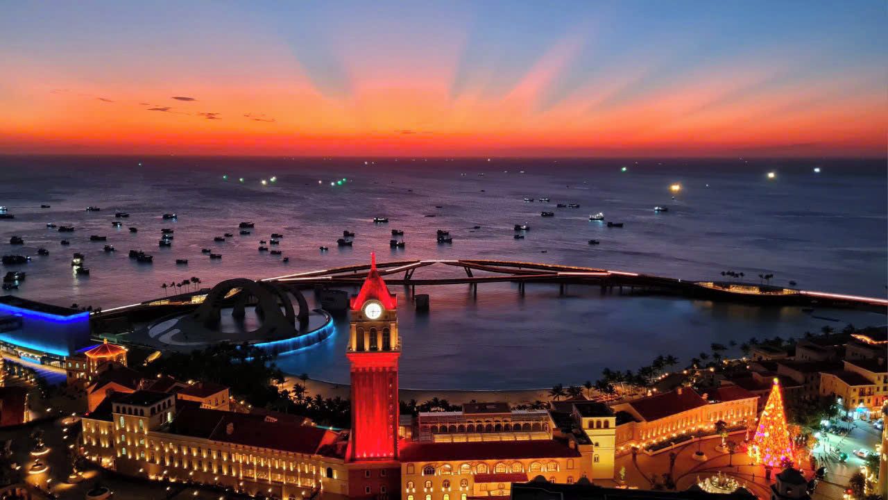 A coastal cityscape at dusk with a prominent clock tower, a marina with boats, and illuminated buildings.