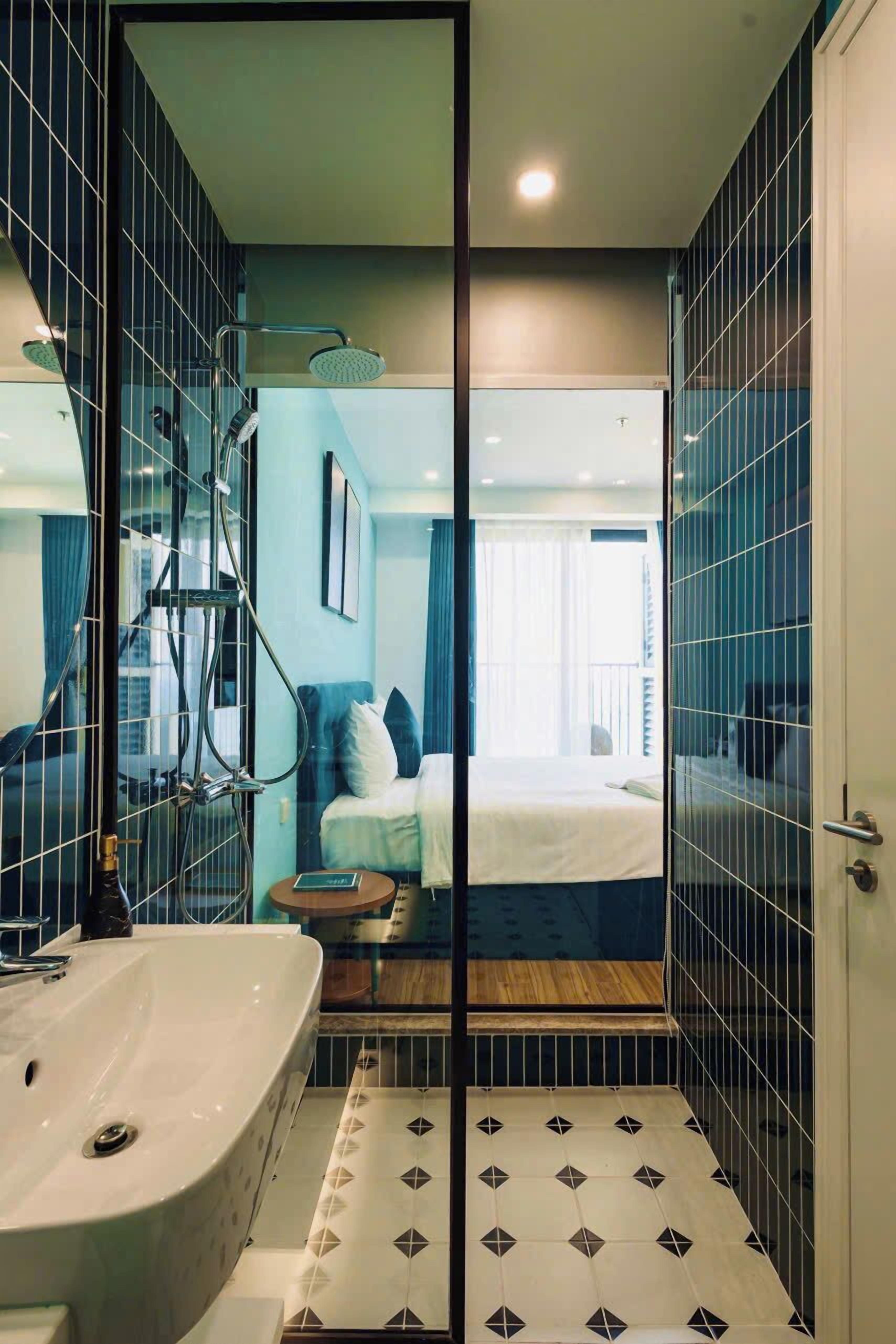 A modern bathroom with a large sink, a shower with a glass door, and a visible bedroom with a bed and curtains.