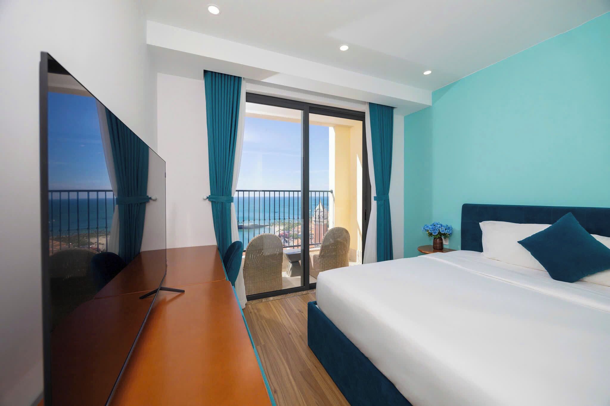 A hotel room with a bed, a TV, a balcony with a view of the sea, and a dining area.