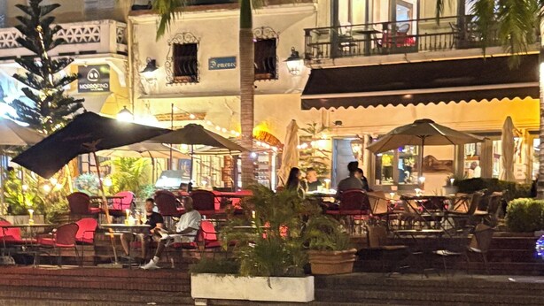 Ein Straßencafé mit Tischen und Stühlen, Palmen und ein Gebäude mit Balkonen im Hintergrund.