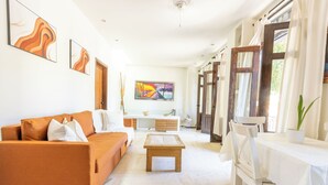 Ein geräumiges Wohnzimmer mit einer Couch, einem Couchtisch und einem Esstisch. An den Wänden hängen Bilder und ein Fenster mit Vorhängen.