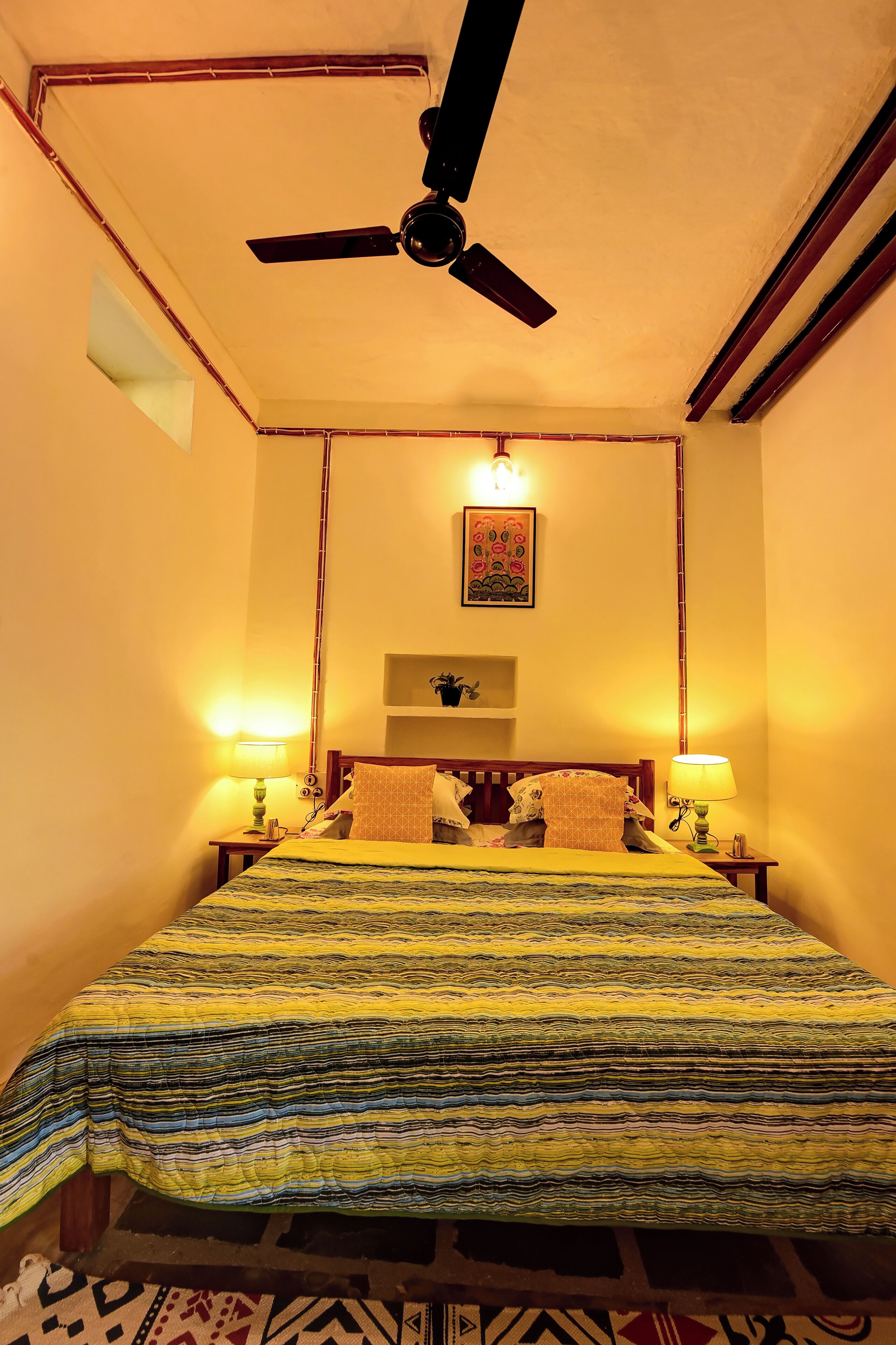 Ein Hotelzimmer mit einem Bett, zwei Nachttischlampen, einem Deckenventilator, einem Wandregal und einem Wandbild.