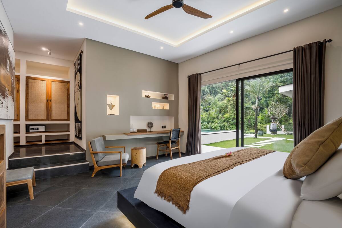 A modern bedroom with a large bed, a desk, and a chair. There is a view of greenery outside the window.