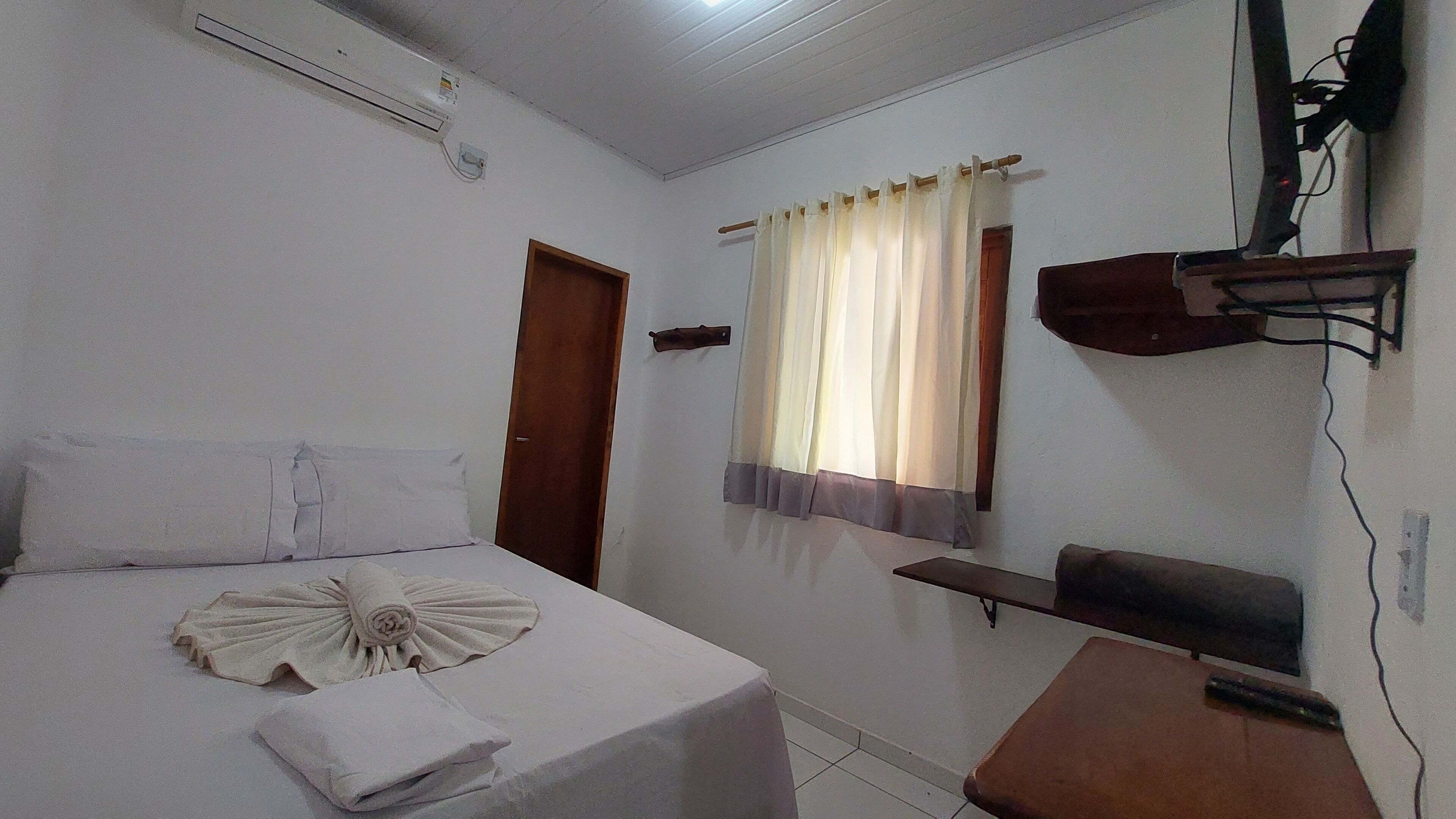 Habitación de hotel con cama, una mesita, televisor montado en la pared y ventana con cortinas.